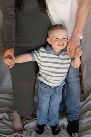 Happy toddler boy in striped shirt and jeans standing between parents during maternity photo session.