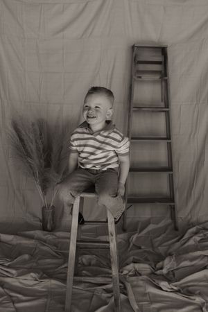Black and white photo of laughing toddler boy sitting on stool near decorative ladder.