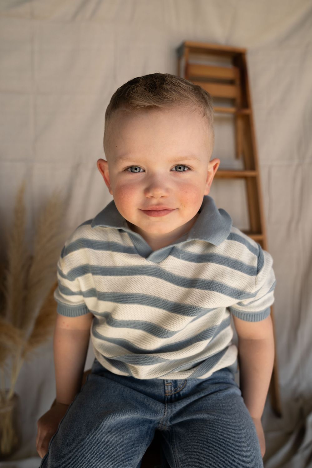 Smiling toddler boy in blue striped polo shirt and jeans sitting in a studio with neutral backdrop.