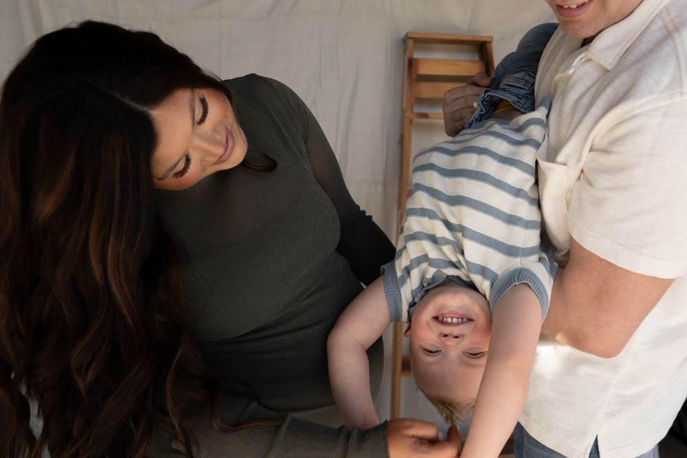 Parents laughing and playing with their happy toddler boy during a cozy family photo session indoors.