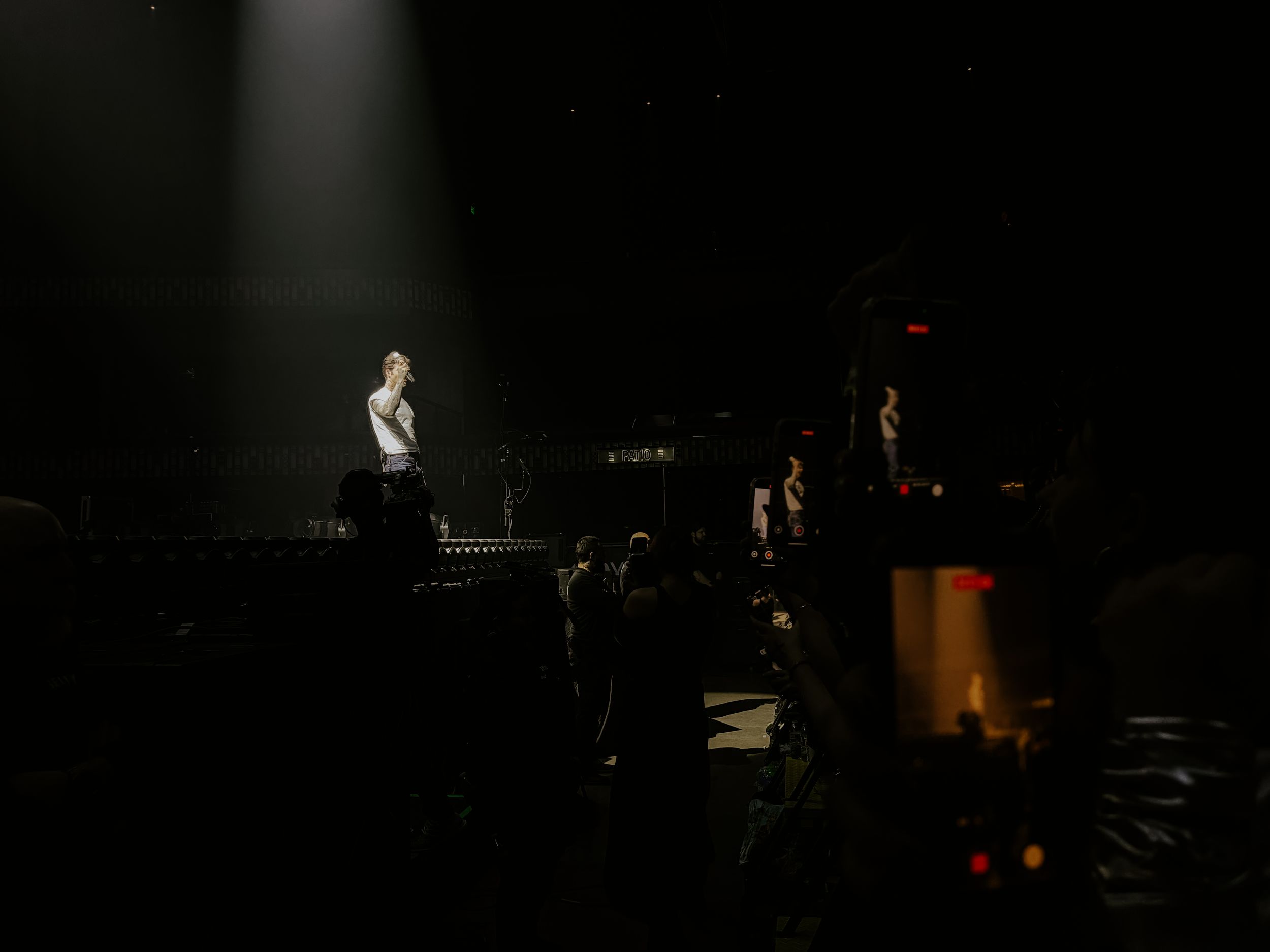 A lone performer stands under a spotlight on a dark concert stage with band members visible in the background.