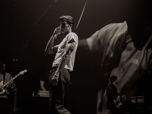 Black and white concert photo of a male singer performing on stage with guitarist in background.