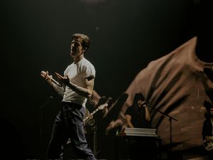 A guitarist performs on a dimly lit stage, wearing a white t-shirt, with a keyboardist visible in the background.