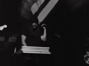 Black and white photo of a long-haired musician playing keyboard on stage during a live performance.