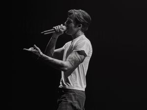 Black and white photo of a tattooed male singer performing on stage, holding a microphone close to his mouth.