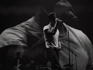 Black and white photo of a male singer performing on stage with a microphone, large projection behind him.