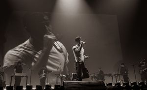 Black and white concert photo of a male performer singing on stage with a large projected image behind him.