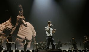 A performer sings on stage with dramatic lighting and a large projected image behind them at a live concert.