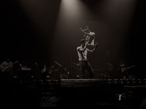 Black and white concert photo of two performers on a dramatic spotlight-lit stage with a full band performing behind them.