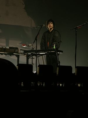 Young musician stands behind keyboard setup on a darkly lit concert stage.