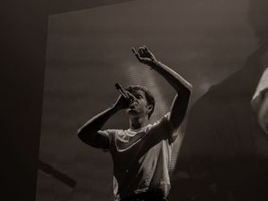 Black and white close-up of a male vocalist singing passionately into a microphone with one arm raised at a live concert.