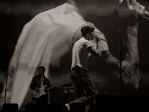 A dynamic black and white photo of a male singer performing on stage with a guitarist behind him.