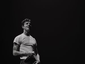 Black and white photo of a male performer on stage in a white t-shirt, looking upward dramatically.