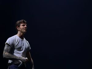 Tattooed male performer on dark stage holding microphone in white t-shirt under dramatic spotlight lighting.