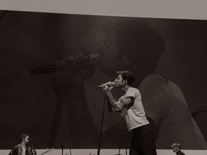 Tattooed singer performs on stage with dramatic shadow projection backdrop in black and white.