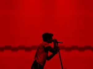 A silhouetted male singer performs passionately into a microphone against a dramatic red-lit stage backdrop.