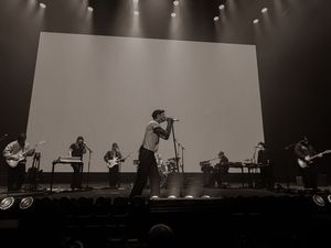 Black and white concert photo of a full band performing on stage with dramatic lighting and large screen.