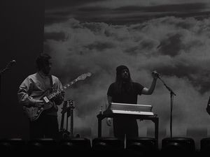 Guitarist and keyboardist perform on stage with clouds projected on backdrop in black and white.