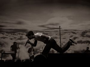 Black and white photo of a performer leaning toward crowd on stage with dramatic cloud backdrop.