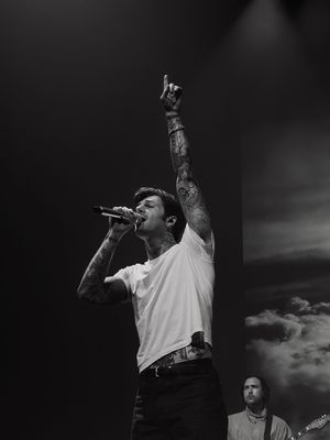 Tattooed male singer performing on stage, raising one arm high while singing into a microphone in black and white.