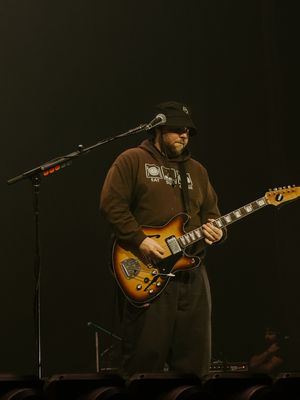 Guitarist in brown hoodie and beanie plays sunburst electric guitar on stage.