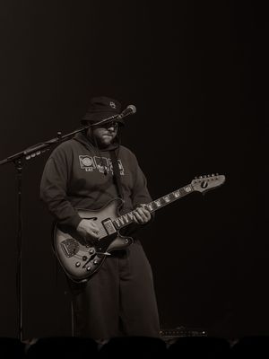 Musician playing electric guitar on stage wearing a hoodie and sunglasses in a moody black and white concert photo.