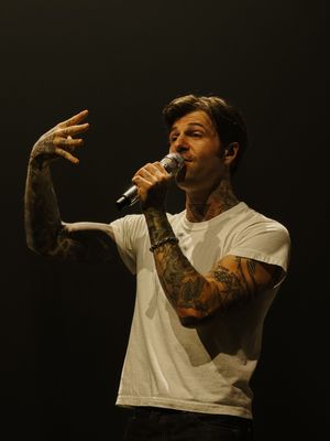 Heavily tattooed male singer in white t-shirt performs with microphone under dramatic stage lighting.