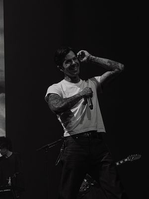 Tattooed male vocalist smiling and performing on stage in a white t-shirt during a live concert in black and white.