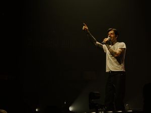 Tattooed male performer in white t-shirt pointing upward on dimly lit concert stage.