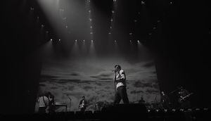 Band performing on stage with dramatic cloud backdrop and concert lighting in black and white.