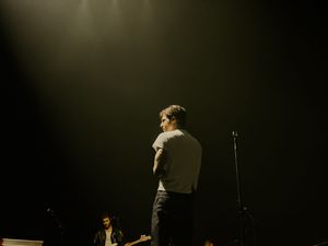Performer standing alone on a dimly lit stage viewed from behind with band members visible in the background.
