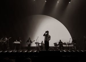 Full band silhouetted against large circular backdrop on concert stage in black and white.