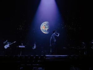 Band performing on dark stage with stunning Earth projection backdrop and single spotlight.