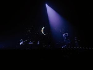 Band performing on dark stage with dramatic crescent moon backdrop and single spotlight beam.