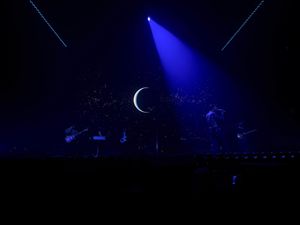 Band performing under blue crescent moon stage backdrop with dramatic laser lighting effects.