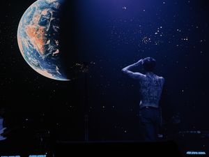 A tattooed performer stands on stage facing a large projected Earth globe against a starry night sky backdrop.