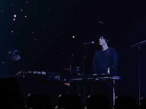 Curly-haired musician at synthesizer keyboard performing on dark stage with starry backdrop.