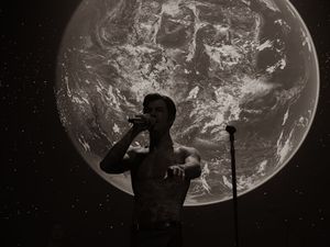 Shirtless performer sings into microphone with giant Earth projection in black and white.