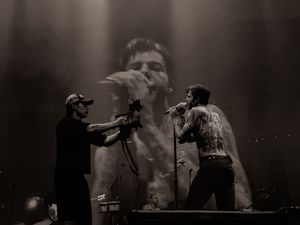 Two performers on stage with large projected face in black and white concert photo.