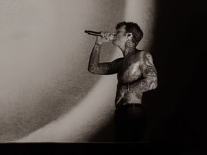 Shirtless tattooed male vocalist sings into microphone against dramatic light in black and white.