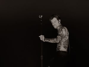 Shirtless tattooed performer grips microphone stand on dimly lit stage in black and white photo.