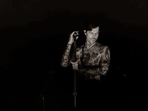 Elegant performer in patterned suit adjusts hat at microphone in moody black and white photo.