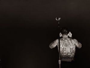 Back view of heavily tattooed shirtless performer at microphone stand in black and white.