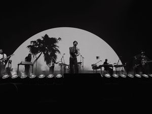 Black and white concert photo of a band performing on stage with a large circular backdrop and palm tree silhouette.