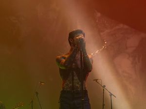 Tattooed shirtless performer covering face with hands in warm orange stage lighting during concert.