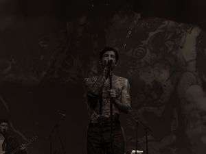 Tattooed vocalist singing into microphone against an ornate illustrated backdrop.