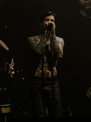 Heavily tattooed shirtless male vocalist singing into microphone on dark concert stage.