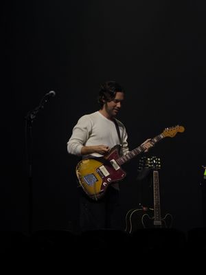 Male guitarist playing vintage Fender guitar on dimly lit concert stage in cream sweater.