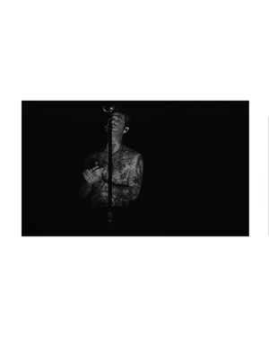 Heavily tattooed male singer performing at microphone in dramatic black and white concert photo.