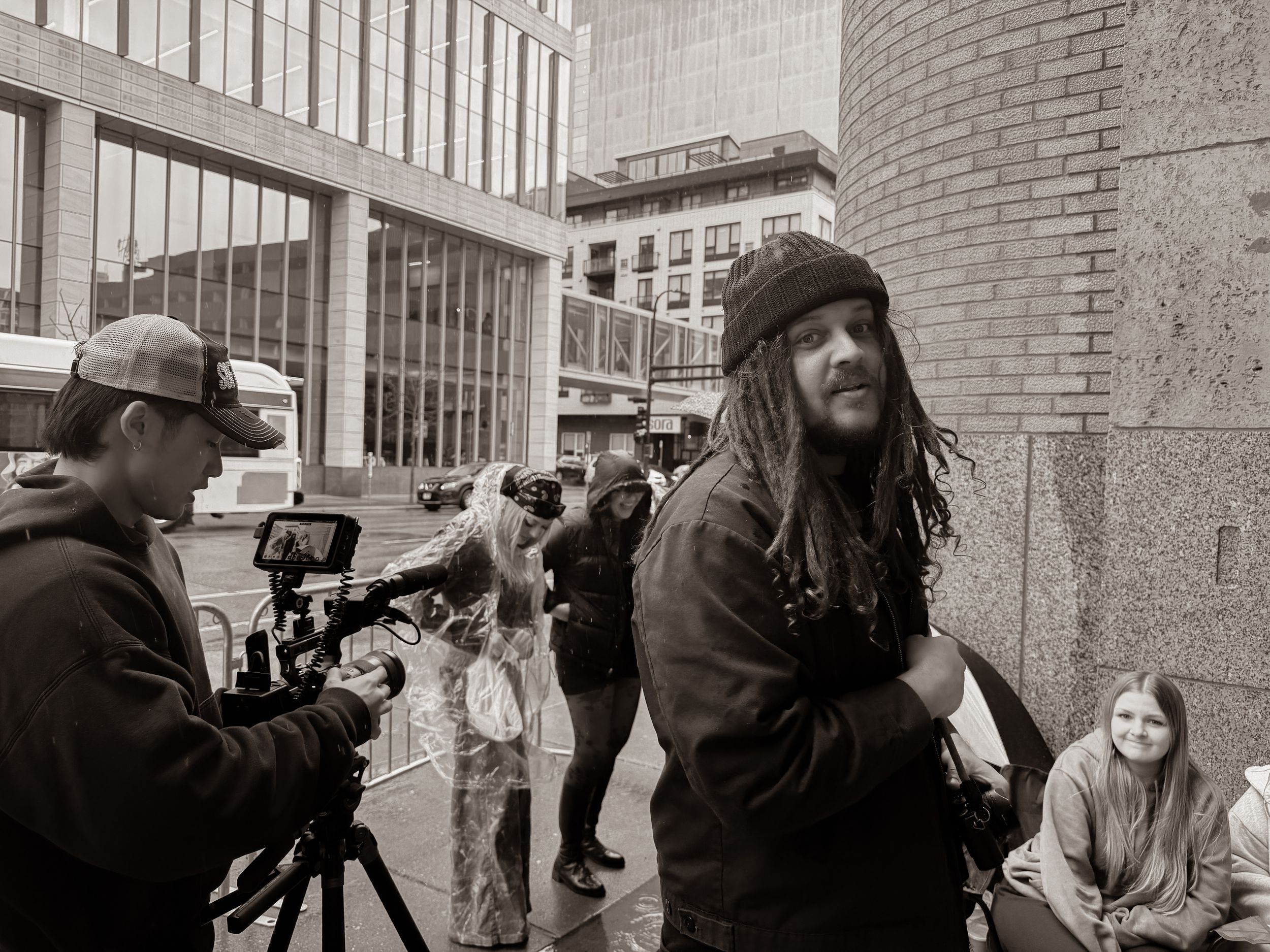 Man with dreadlocks stands near film camera crew on a rainy city street in black and white.